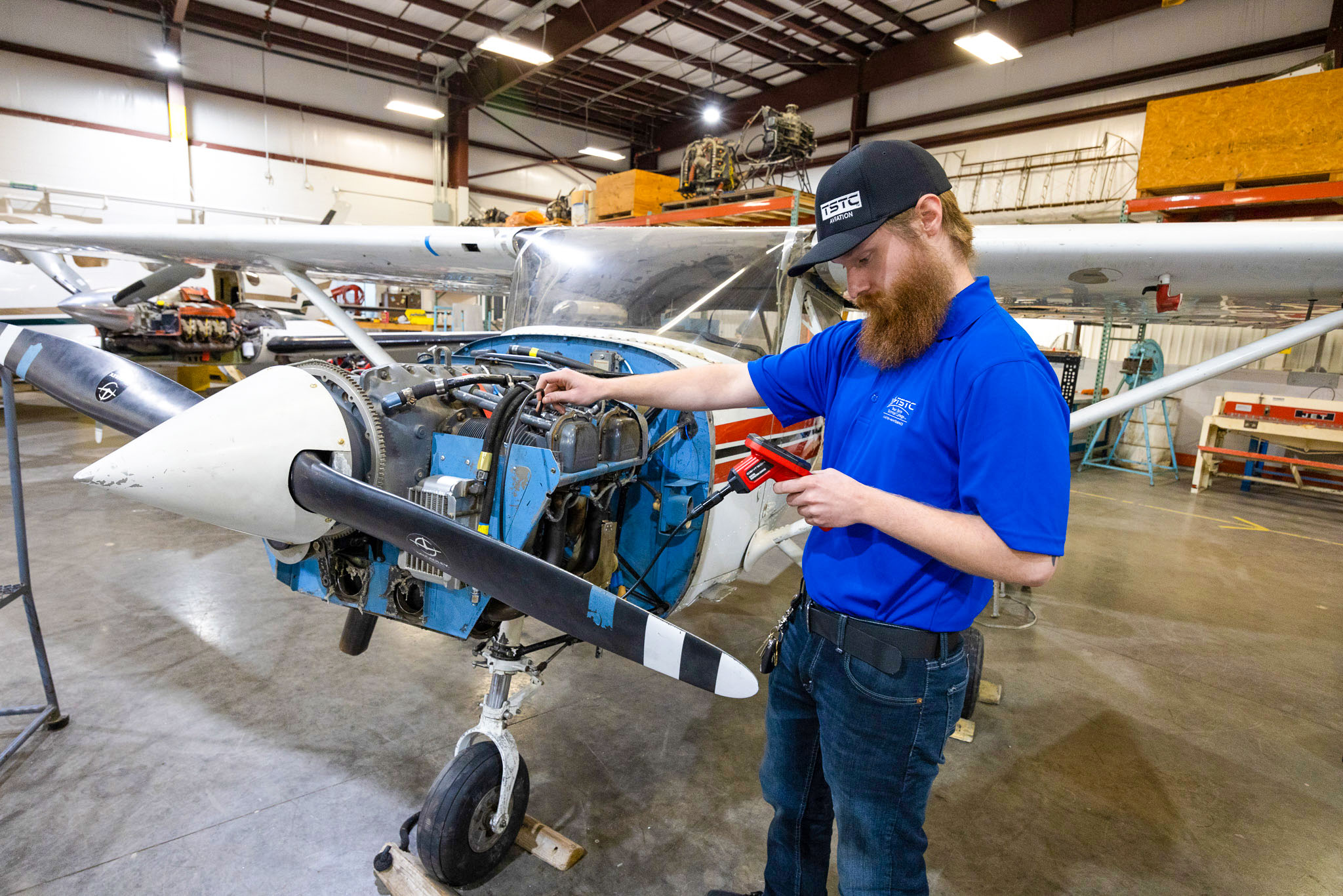 Student in a blue shirt and cap uses a handheld diagnostic tool to inspect wiring on an exposed aircraft engine mounted on a small propeller plane inside a hangar-like workshop.